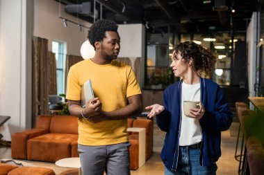 Elinde kahve tutan kadın ve not defterli adam birlikte çalışma ortamında sohbet ediyorlar.
