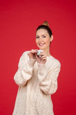 Yüz bakımı. Kırmızı arka planda elinde krema kavanozuyla güzel bir genç kadın.