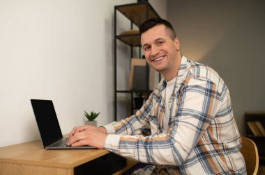 Ev ofisinin ahşap masasında dizüstü bilgisayarda çalışan mutlu adam.