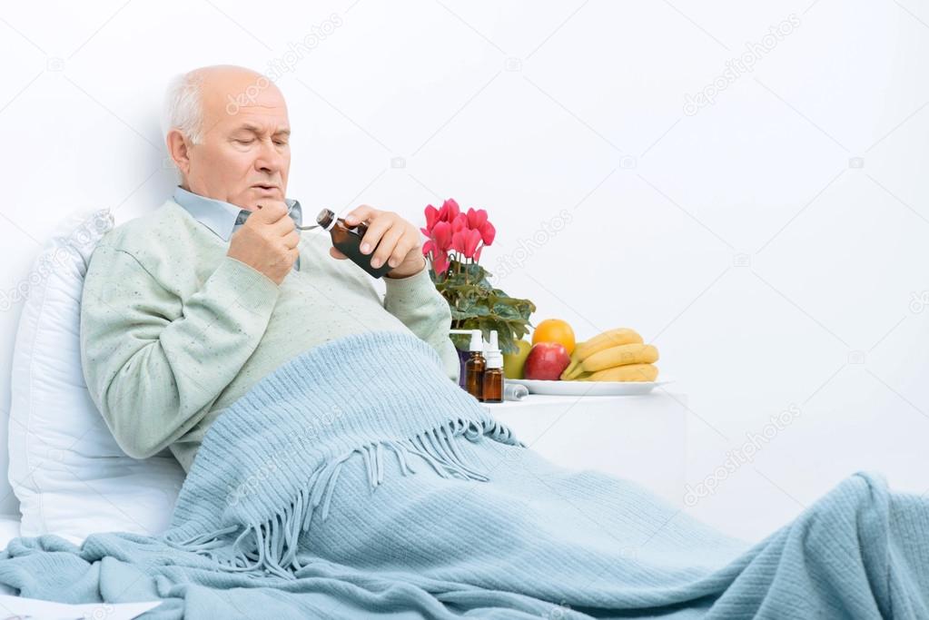 Elderly man rests in bed and pours cough syrup.