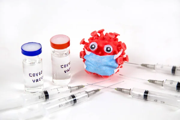 Beyaz bir zemin üzerinde covid-19 aşısı olan tıbbi şırınga ve şişelerle çevrili, kabarık gözlü ve mavi maskeli plastik bir kırmızı top. İlaç viral enfeksiyon salgını aşıları