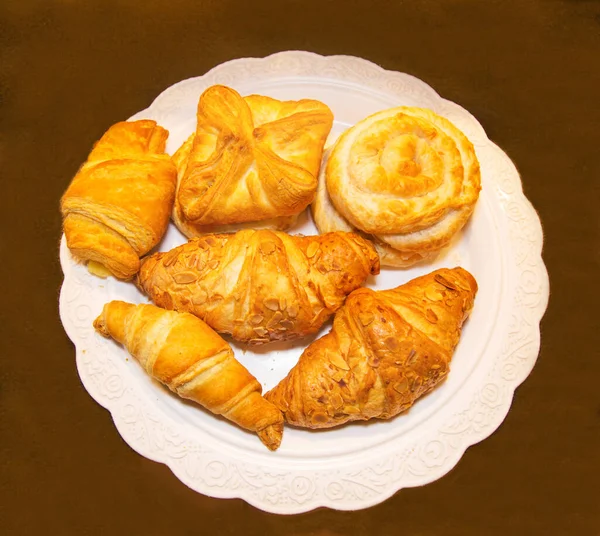 Pastry made of puff pastry in the form of round twisted buns with jam, coconut chips, croissants on an openwork white napkin of a round shape. Confectionery products for tea and coffee.