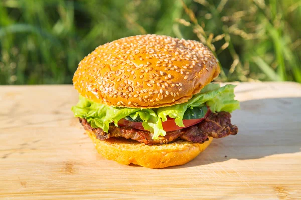 肉ステーキ トマト キュウリ レタスの黄色のハンバーガーは 晴れた日にまな板と緑の草を背景にしています バーベキュー製品の調理 — ストック写真