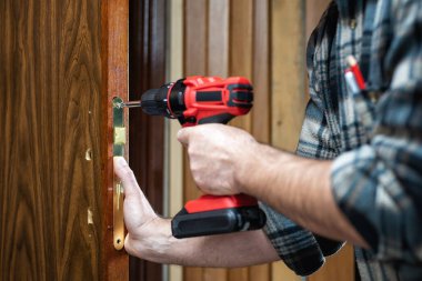 Yakın plan. Elektrikli tornavidayla marangoz ahşap bir kapının kilidini tamir ediyor. İnşaat endüstrisi. 