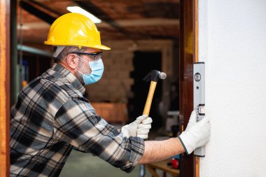 Carpenter worker at work repairs and installs a room door, wear the surgical mask to prevent Coronavirus infection. Preventing Pandemic Covid-19 at the workplace. Carpentry. clipart
