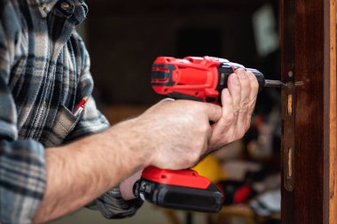 Yakın plan. Elektrikli tornavidayla marangoz ahşap kapının kilidi için plaketi tamir ediyor. İnşaat endüstrisi. 