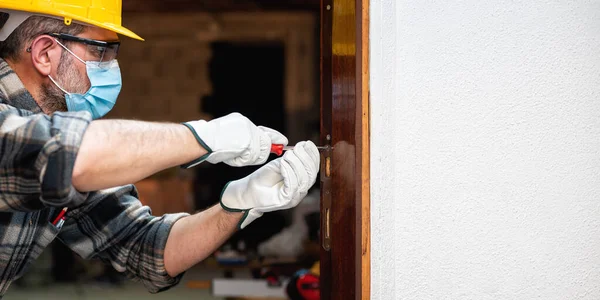 Carpenter worker at work repairs and installs a room door, wear the surgical mask to prevent Coronavirus infection. Preventing Pandemic Covid-19 at the workplace. Carpentry. Text space.