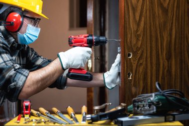 Marangoz, Coronavirus enfeksiyonunu önlemek için bir oda kapısı kilidi takıp cerrahi maske takıyor. İş yerinde Covid-19 salgınını önlemek. Marangozluk.