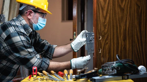 Carpenter worker at work repair and install a room door lock, wear the surgical mask to prevent Coronavirus infection. Preventing Pandemic Covid-19 at the workplace. Carpentry.