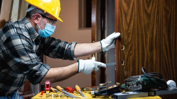 Carpenter worker at work repair and install a room door lock, wear the surgical mask to prevent Coronavirus infection. Preventing Pandemic Covid-19 at the workplace. Carpentry.