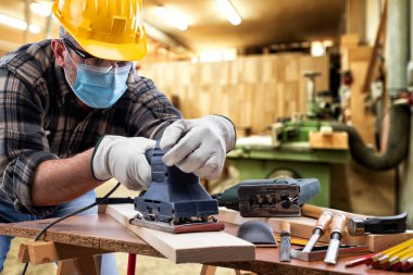 Marangozluk atölyesinde çalışan marangoz, Coronavirus enfeksiyonunu önlemek için kask, gözlük, deri eldiven ve cerrahi maske takıyor. İş yerinde salgının önlenmesi Covid-19.