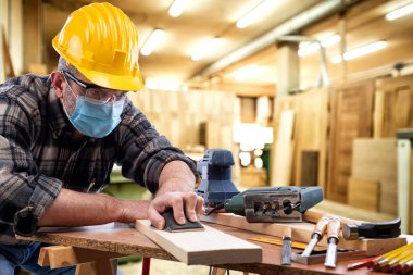 Marangozluk atölyesinde çalışan marangoz, Coronavirus enfeksiyonunu önlemek için kask, gözlük ve cerrahi maske takıyor. İş yerinde salgının önlenmesi Covid-19.