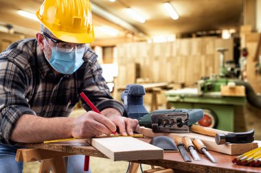 Marangozluk atölyesinde çalışan marangoz, Coronavirus enfeksiyonunu önlemek için kask, gözlük ve cerrahi maske takıyor. İş yerinde salgının önlenmesi Covid-19.