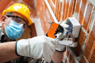 Kablo sökücü penseyle elektrik işçisi elektrik kablolarını hazırlar, Coronavirus 'un yayılmasını önlemek için cerrahi maske takar. Covid-19, iş güvenliği; inşaat endüstrisi.