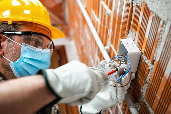 Tornavidalı bir elektrikçi elektrik sisteminin düğmesindeki kabloyu tamir eder, Coronavirus 'un yayılmasını önlemek için cerrahi maske takar. Covid-19, iş güvenliği; inşaat endüstrisi. 