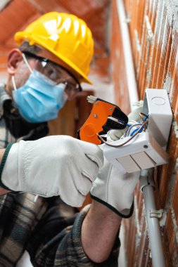 Kablo sökücü penseyle elektrik işçisi elektrik kablolarını hazırlar, Coronavirus 'un yayılmasını önlemek için cerrahi maske takar. Covid-19, iş güvenliği; inşaat endüstrisi.