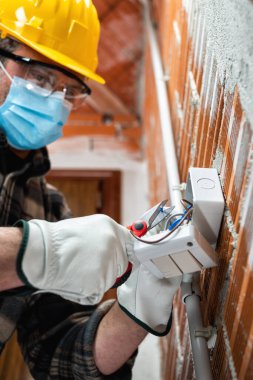 Elektrik işçisi elinde makasla bir elektrik sisteminin elektrik kablolarını hazırlar; Coronavirus 'un yayılmasını önlemek için cerrahi maske takar. Covid-19, iş güvenliği; inşaat endüstrisi.