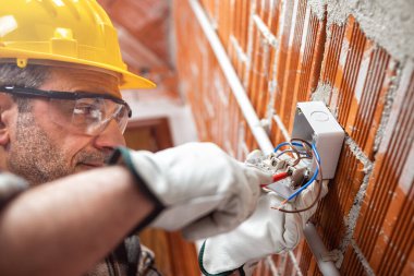 Tornavidayla çalışan bir elektrik işçisi elektrik sisteminin düğmesindeki kabloyu tamir eder; kask, eldiven ve gözlük takar. İşyeri güvenliği. 