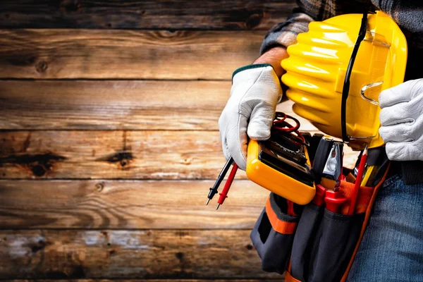 Electrician worker on vintage wooden background; holds the multimeter tester, helmet and protective goggles in hand. Construction industry, electrical system.