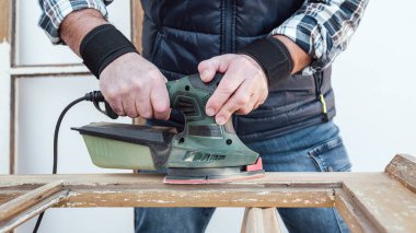 Zanaatkar. Yetişkin bir marangoz eski bir ahşap pencereyi düzeltmek için elektrikli zımpara makinesi kullanıyor. İnşaat endüstrisi, marangozluk, ev işi. Yenileme.