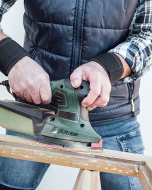 Zanaatkar. Yetişkin bir marangoz eski bir ahşap pencereyi düzeltmek için elektrikli zımpara makinesi kullanıyor. İnşaat endüstrisi, marangozluk, ev işi. Yenileme.