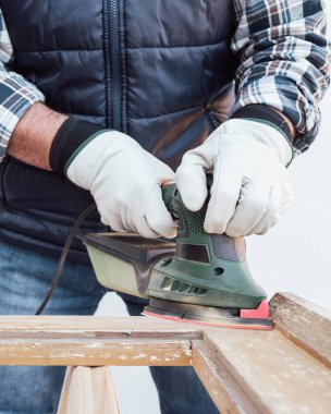 Zanaatkar. Yetişkin bir marangoz eski bir ahşap pencereyi düzeltmek için elektrikli zımpara makinesi kullanıyor. İnşaat endüstrisi, marangozluk, ev işi kendin yap, işyeri güvenliği. Yenileme.