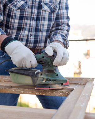 Zanaatkar. Yetişkin bir marangoz eski bir ahşap pencereyi düzeltmek için elektrikli zımpara makinesi kullanıyor. İnşaat endüstrisi, marangozluk, ev işi kendin yap, işyeri güvenliği. Yenileme.