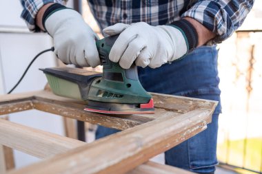 Zanaatkar. Yetişkin bir marangoz eski bir ahşap pencereyi düzeltmek için elektrikli zımpara makinesi kullanıyor. İnşaat endüstrisi, marangozluk, ev işi kendin yap, işyeri güvenliği. Yenileme.