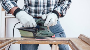 Zanaatkar. Yetişkin bir marangoz eski bir ahşap pencereyi düzeltmek için elektrikli zımpara makinesi kullanıyor. İnşaat endüstrisi, marangozluk, ev işi. Kendin yap. İş yeri güvenliği. Yenileme.