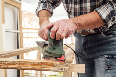 Zanaatkar. Yetişkin bir marangoz eski bir ahşap pencereyi düzeltmek için elektrikli zımpara makinesi kullanıyor. İnşaat endüstrisi, marangozluk, ev işi. Yenileme.