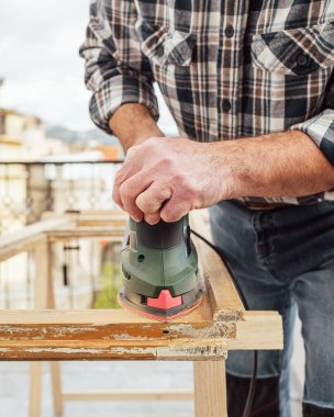 Zanaatkar. Yetişkin bir marangoz eski bir ahşap pencereyi düzeltmek için elektrikli zımpara makinesi kullanıyor. İnşaat endüstrisi, marangozluk, ev işi. Kendin yap. Yenileme.