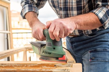 Zanaatkar. Yetişkin bir marangoz eski bir ahşap pencereyi düzeltmek için elektrikli zımpara makinesi kullanıyor. İnşaat endüstrisi, marangozluk, ev işi. Kendin yap. Yenileme.