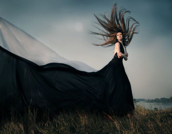 Fashion female model with long blowing hair outdoor. Portrait of sexy glamour beautiful woman with healthy and beauty flying fluttering long brown hair wearing black dress resists strong wind. Copy space.