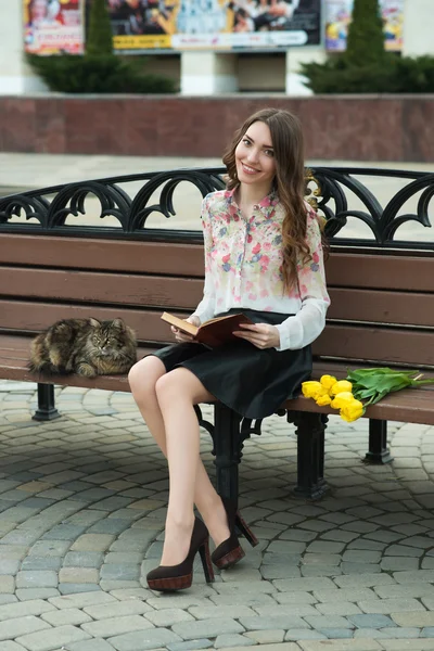 Chica leyendo un libro con un gato en un banco en la ciudad — Foto de Stock