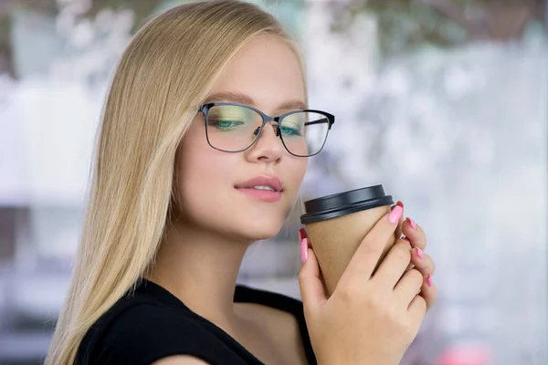 Beautiful young blonde woman with perfect skin, natural long light blond hair and glasses stands front of the window. Portrait of beauty model teen girl with craft paper cup of take-off coffee.