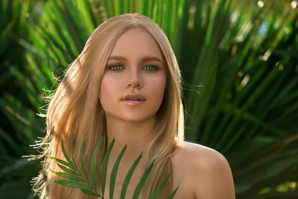 Beautiful young blonde woman with perfect skin and long light blond hair front of plant tropical green leaves background. SPA, wellness, bodycare and skincare.  Tropical background, selective focus.