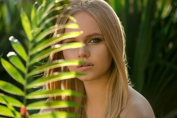 Beautiful young blonde woman with perfect skin and long light blond hair front of plant tropical green leaves background. SPA, wellness, bodycare and skincare.  Tropical background, selective focus.
