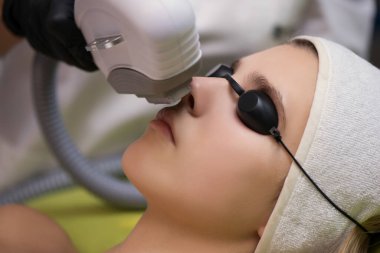 Terapist güzellik uzmanı güzellik merkezindeki genç kadının yüzüne lazer tedavisi uyguluyor. Yüz lazeri epilasyon prosedürleri. Kuafördeki cihazlarla modeller için yüz bakımı yenileme tıbbi düzeltme. Yaklaş, seçici odaklanma.