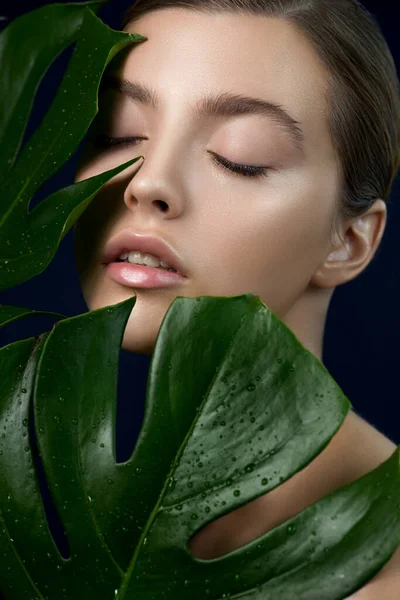Portrait of beautiful young woman with perfect skin posing with closed eyes through the tropical green leaves. Attractive lady enjoys her skin care procedures. SPA, wellness and skincare concept.