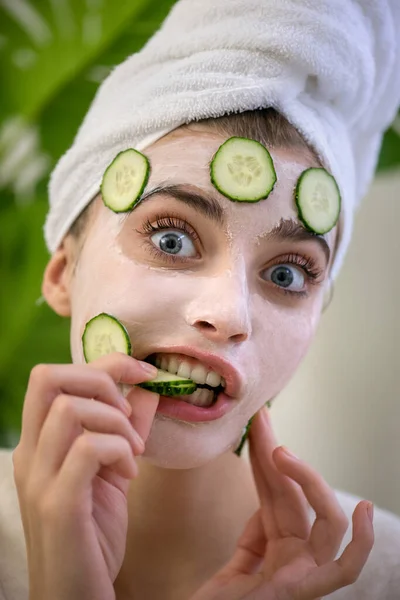Young woman applies face mask with fresh cucumbers on her face and makes makes a funny face. Beauty model with  perfect fresh skin and long eyelashes cares about her skin at home. Close up, selected focus.
