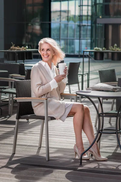 Attractive elderly woman with gray hair in casual clothes with cup of coffee sits in the cafe. Freelance business woman enjoys her hot beverage in the open space.