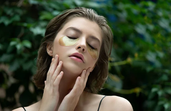 Beautiful young woman with closed eyes enjoyed green  and gold eye mask fron of green tropical background. Portrait of beauty model with natural  makeup cares about her skin. Spa, skincare and wellness. Selective focus.