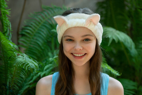 Beautiful little teenage girl with funny cat headband is smiling front of tropical leaves background. Portrait of cute little child enjoying skin care procedures.