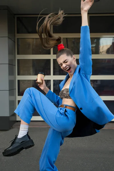 Beautiful young woman in a blue suit with a paper cup of takeaway coffee is dancing on a city street. Fashion model with a cup of coffee. Success, achievement, victory concept.