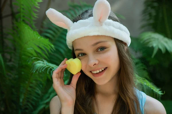可爱的小女孩享受皮肤护理程序的肖像 漂亮的小女孩 戴着滑稽的兔子头带 用心形海绵擦拭她的脸 图库图片