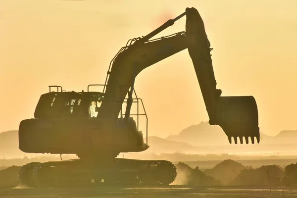 An excavating machine is moving earth in a very dusty desert environment in the morning sunrise hours.