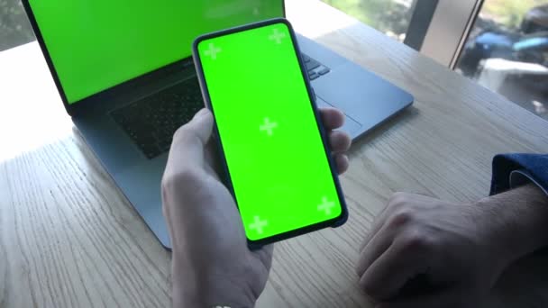 Макет зеленого екрану смартфона в руках чоловіка на тлі навчальної роботи ноутбука онлайн — стокове відео
