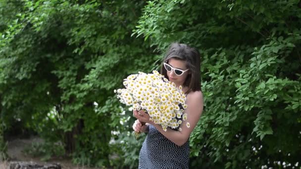Jong meisje in glazen met een boeket witte madeliefjes staat op straat en ruikt een boeket bloemen — Stockvideo