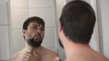 Banyodaki aynaya yansıyan adam yansımaya bakar ve sakalına dokunur.