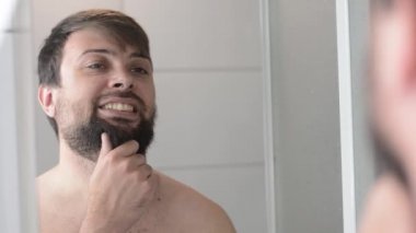 Banyodaki aynaya yansıyan adam yansımaya bakar ve sakalına dokunur.